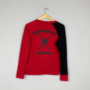 May include: Red sweatshirt with a black sleeve. The back features a skull and crossbones graphic with the words "NOISE FREAK OUT" and "MUSICA HOLIC" in gray lettering. The sweatshirt is hanging on a wooden hanger.