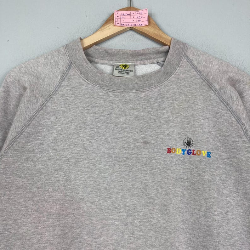May include: A light gray sweatshirt with the "BODY GLOVE" logo in rainbow colors on the front. The sweatshirt has a crew neck and raglan sleeves. The Body Glove logo is a hand inside a circle.
