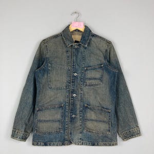 May include: A vintage-style denim jacket in faded blue, featuring a button-up closure, four front pockets, and a classic collar. The jacket has a distressed look, suggesting it has been pre-worn. It is displayed on a hanger against a neutral backdrop.