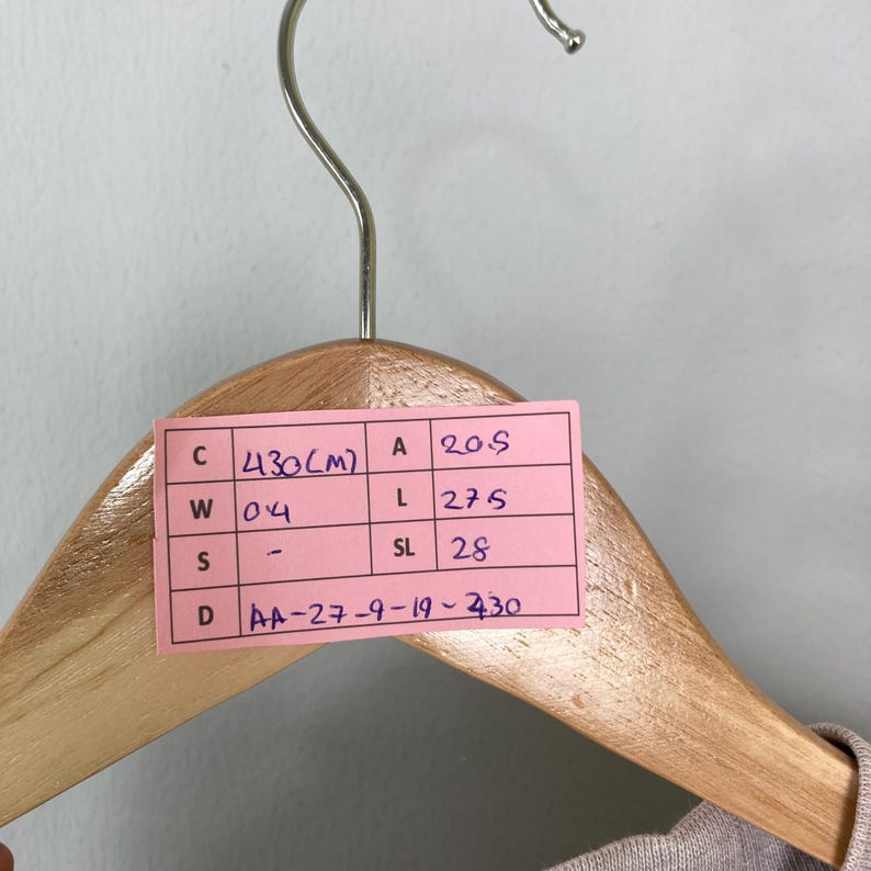 May include: A close-up of a wooden clothes hanger with a pink tag attached. The tag has handwritten text and measurements. The hanger is suspended against a light gray background.