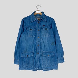 K&ouml;nnte beinhalten: Blaue Jeansjacke mit vier Vordertaschen und Knopfverschluss. Die Jacke hat einen Kragen und lange &Auml;rmel. Das Kleidungsst&uuml;ck h&auml;ngt an einem Holzb&uuml;gel vor einem hellgrauen Hintergrund.