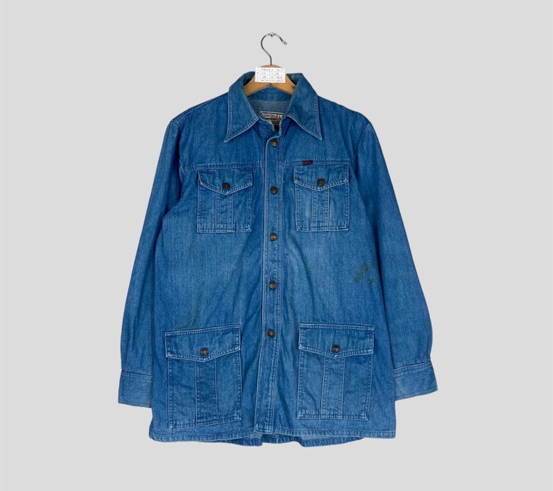 K&ouml;nnte beinhalten: Blaue Jeansjacke mit vier Vordertaschen und Knopfverschluss. Die Jacke hat einen Kragen und lange &Auml;rmel. Das Kleidungsst&uuml;ck h&auml;ngt an einem Holzb&uuml;gel vor einem hellgrauen Hintergrund.