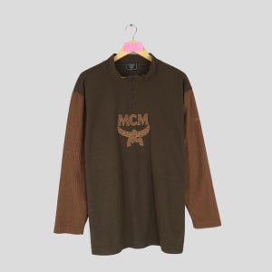 Puede incluir: Un jersey marrón de manga larga con cuello con cremallera. Las mangas son de color marrón acanalado y el pecho presenta el logotipo de MCM en un marrón más claro. El jersey está colgado de una percha.