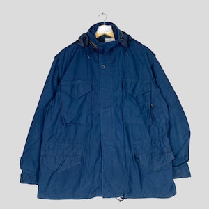 Puede incluir: Chaqueta azul marino de estilo militar con capucha, múltiples bolsillos y cierre de botones. La chaqueta está colgada de una percha sobre un fondo gris claro. La chaqueta tiene un cordón en la parte inferior.