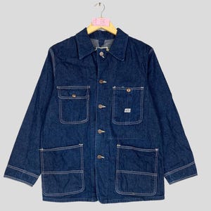 May include: A dark blue denim jacket with four front pockets and button fastenings. The jacket features a classic collar and long sleeves. White stitching outlines the pockets and seams. The jacket is hanging on a wooden hanger.