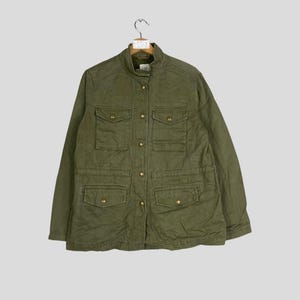 Könnte beinhalten: Olivgrüne Jacke mit vier Fronttaschen und goldfarbenen Knöpfen. Die Jacke hat einen Stehkragen und hängt an einem Holzbügel. Die Jacke ist aus strapazierfähigem Stoff gefertigt und für verschiedene Wetterbedingungen geeignet.