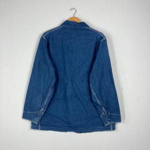 Puede incluir: Chaqueta vaquera azul con mangas largas, pu&ntilde;os abotonados y cuello. La chaqueta est&aacute; colgada de una percha de madera sobre un fondo blanco. Las costuras son visibles a lo largo de las costuras.