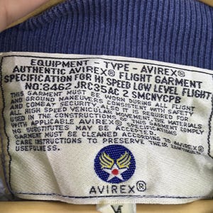 Puede incluir: Primer plano de una etiqueta de una prenda de vuelo Avirex. La etiqueta blanquecina presenta texto negro que detalla las especificaciones y el nombre de la marca. Tambi&eacute;n es visible un emblema azul y amarillo.