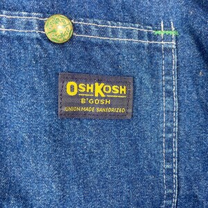 Puede incluir: Primer plano de tela vaquera azul. Un bot&oacute;n verde en la parte superior izquierda. Una etiqueta azul marino con texto amarillo dice "OSH KOSH B'GOSH UNION MADE SANFORIZED". Costuras blancas y detalles verdes.