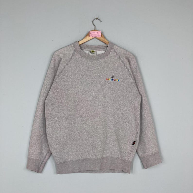 May include: A heather gray sweatshirt with the embroidered text "BODY GLOVE" in rainbow colors. The long-sleeved crew neck sweatshirt has a ribbed collar, cuffs, and hem. A small black tag is sewn on the lower left side.