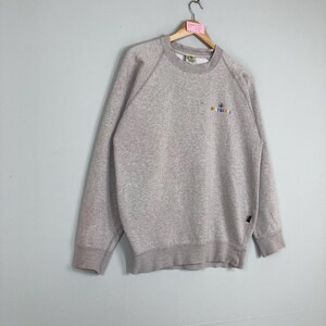 May include: A light gray sweatshirt with long sleeves and a crew neck. The sweatshirt has a small, colorful logo that reads "RECYCLE" in a rainbow font. The garment is hanging on a wooden hanger.