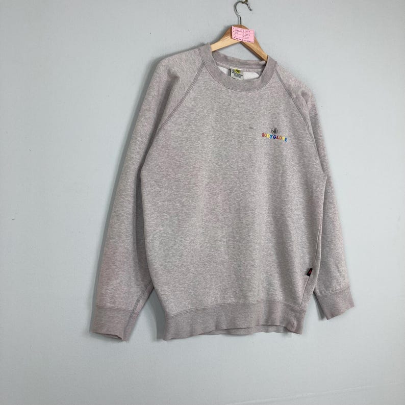 May include: A light gray sweatshirt with long sleeves and a crew neck. The sweatshirt has a small, colorful logo that reads "RECYCLE" in a rainbow font. The garment is hanging on a wooden hanger.