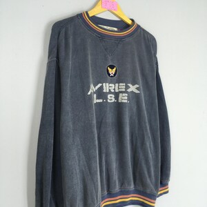 Puede incluir: Una sudadera azul oscuro con el texto "AVIREX L.S.E." en blanco. La sudadera tiene cuello redondo con un dise&ntilde;o a rayas en amarillo, naranja y azul. El mismo dise&ntilde;o a rayas est&aacute; en los pu&ntilde;os y el dobladillo inferior. Un peque&ntilde;o parche est&aacute; en el pecho.