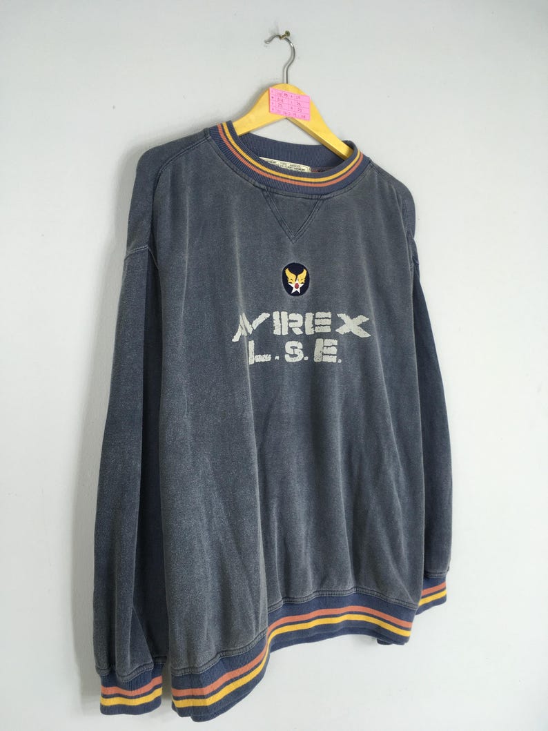 Puede incluir: Una sudadera azul oscuro con el texto "AVIREX L.S.E." en blanco. La sudadera tiene cuello redondo con un dise&ntilde;o a rayas en amarillo, naranja y azul. El mismo dise&ntilde;o a rayas est&aacute; en los pu&ntilde;os y el dobladillo inferior. Un peque&ntilde;o parche est&aacute; en el pecho.