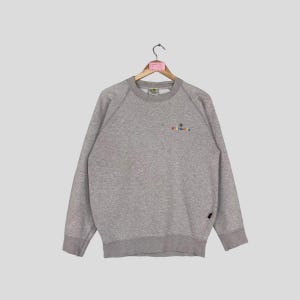 May include: A heather gray sweatshirt with a crew neck and long sleeves. The sweatshirt features a small, colorful embroidered design on the upper left chest, with the text "BODY GLOVE". The garment is hanging on a wooden hanger.
