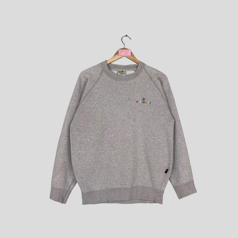 May include: A heather gray sweatshirt with a crew neck and long sleeves. The sweatshirt features a small, colorful embroidered design on the upper left chest, with the text "BODY GLOVE". The garment is hanging on a wooden hanger.