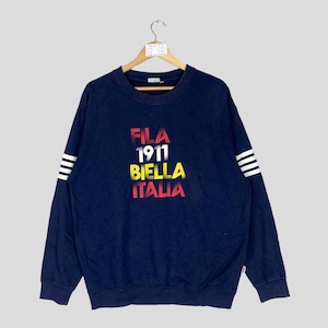 May include: Navy blue sweatshirt with the text "FILA 1911 BIELLA ITALIA" in red, white, yellow, and red. The long sleeves have white stripes near the cuffs. The sweatshirt is hanging on a wooden hanger.