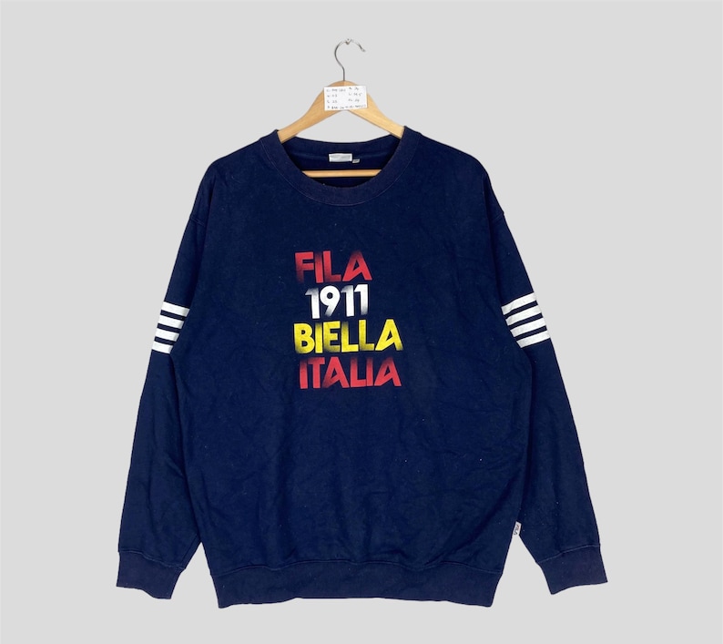 May include: Navy blue sweatshirt with the text "FILA 1911 BIELLA ITALIA" in red, white, yellow, and red. The long sleeves have white stripes near the cuffs. The sweatshirt is hanging on a wooden hanger.