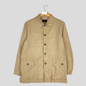 May include: A tan button-down jacket with a collared neckline and two front pockets. The jacket is hanging on a wooden hanger against a light gray background. The jacket has a classic, casual style.