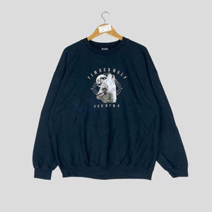 May include: A navy blue sweatshirt with a graphic of a howling wolf under a full moon. The text "TIMBERWOLF COUNTRY" is printed above and below the wolf image in a circular design. The sweatshirt has a crew neck and long sleeves.