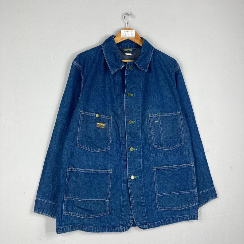 Puede incluir: Chaqueta vaquera azul con cuello, cuatro bolsillos delanteros y botones. La chaqueta tiene un peque&ntilde;o parche con el logo en el pecho y est&aacute; colgada de una percha de madera. El vaquero es de lavado medio.