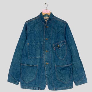 Op de afbeelding: Een blauw spijkerjack met een opstaande kraag, knoopsluiting en meerdere zakken. De jas heeft een klassiek ontwerp met een rood logolabel op een zak. De jas hangt aan een houten hanger.