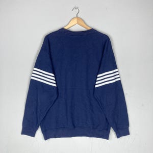 May include: Navy blue sweatshirt with three white stripes on each sleeve. The crew neck sweatshirt is hanging on a wooden hanger against a light gray background. The garment is made of a soft material.