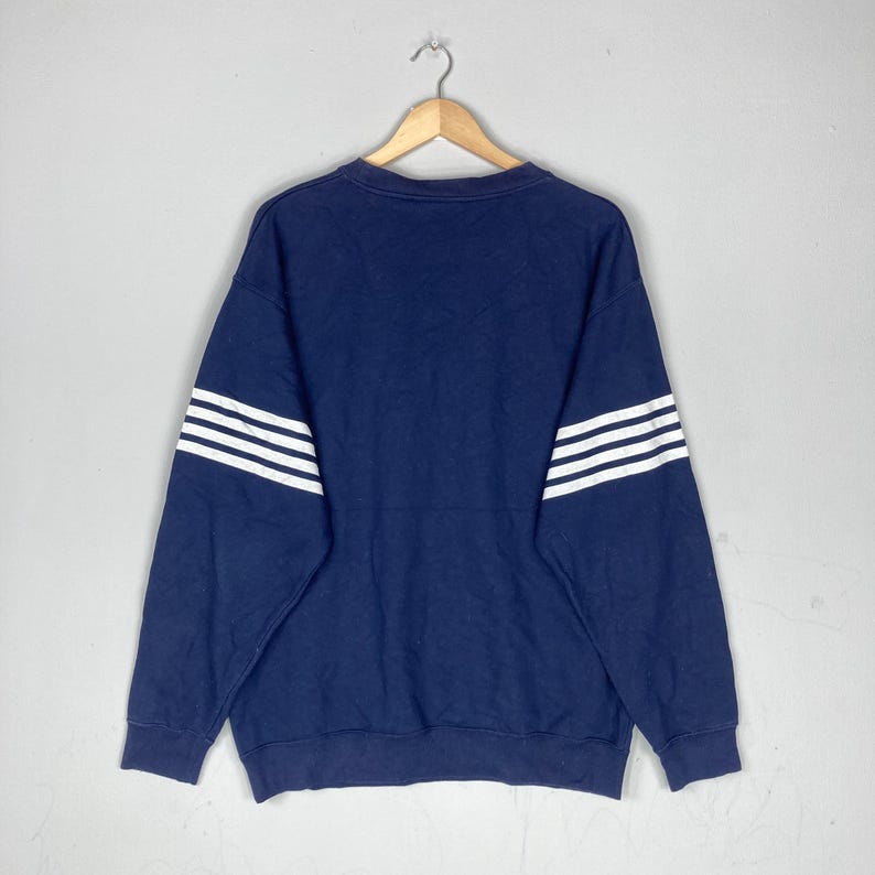 May include: Navy blue sweatshirt with three white stripes on each sleeve. The crew neck sweatshirt is hanging on a wooden hanger against a light gray background. The garment is made of a soft material.