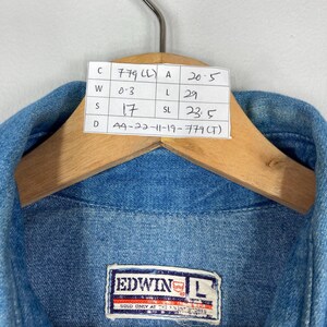 K&ouml;nnte beinhalten: Nahaufnahme einer blauen Jeansjacke mit einem angebrachten Etikett. Das Etikett zeigt Ma&szlig;e und den Markennamen "EDWIN". Die Jacke h&auml;ngt an einem Holzb&uuml;gel. Das Etikett enth&auml;lt den Text "SOLD ONLY AT THE FINEST STORES".