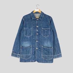 May include: A blue denim jacket with a button-up front, four front pockets, and long sleeves. The jacket is hanging on a wooden hanger against a light gray background. The denim has a faded appearance.