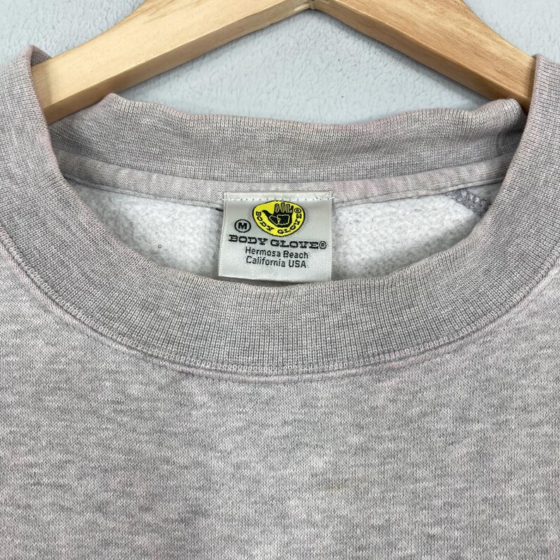 May include: Close-up of a gray sweatshirt with a crew neck. The tag reads "BODY GLOVE Hermosa Beach California USA" with a yellow logo. The sweatshirt is hanging on a wooden hanger.