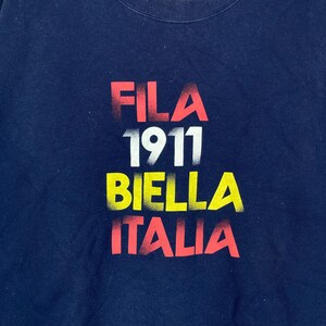 May include: Navy blue sweatshirt with the text "FILA 1911 BIELLA ITALIA" in red, white, and yellow. The text has a distressed, faded appearance. The sweatshirt is a solid color.