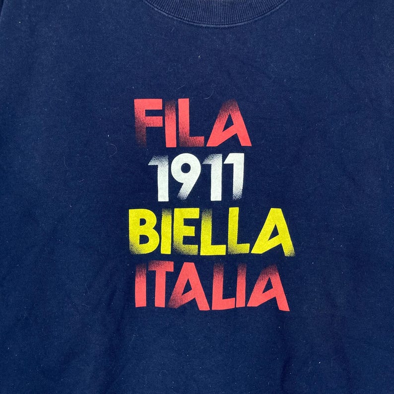 May include: Navy blue sweatshirt with the text "FILA 1911 BIELLA ITALIA" in red, white, and yellow. The text has a distressed, faded appearance. The sweatshirt is a solid color.