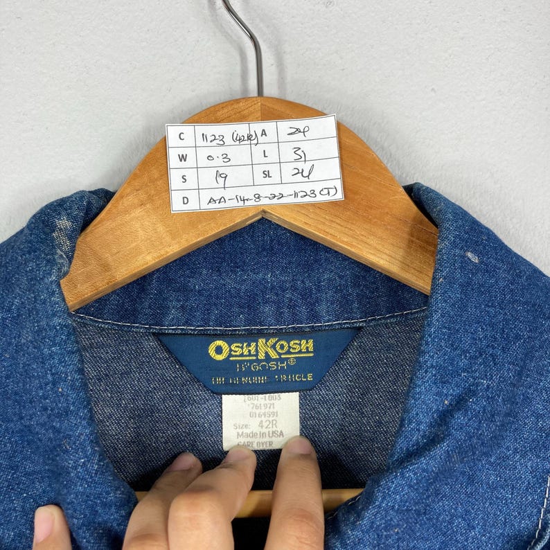Puede incluir: Primer plano de una chaqueta vaquera azul con cuello, colgada de una percha de madera. La chaqueta tiene una etiqueta con la marca "OshKosh" e informaci&oacute;n sobre la talla. Tambi&eacute;n es visible una etiqueta con medidas.