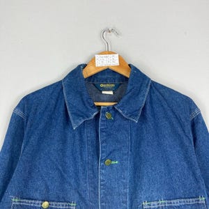 Puede incluir: Chaqueta vaquera azul con cuello y cierre de botones. La chaqueta tiene botones dorados y detalles de costura verdes. El nombre de la marca "OutKast" es visible en la etiqueta.