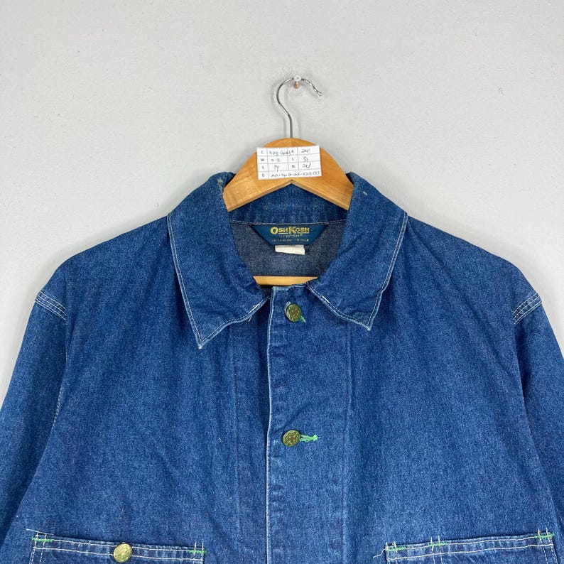 Puede incluir: Chaqueta vaquera azul con cuello y cierre de botones. La chaqueta tiene botones dorados y detalles de costura verdes. El nombre de la marca "OutKast" es visible en la etiqueta.