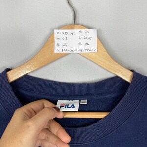 May include: A navy blue FILA sweatshirt hanging on a wooden hanger. The brand logo is visible on the front. A handwritten tag with measurements is attached to the hanger. The sweatshirt is a size 4L.