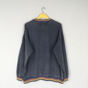 Puede incluir: Una sudadera azul oscuro con cuello redondo y mangas largas, colgada de una percha amarilla. El cuello y el dobladillo presentan rayas amarillas, naranjas y azules. La prenda tiene un aspecto descolorido y vintage.