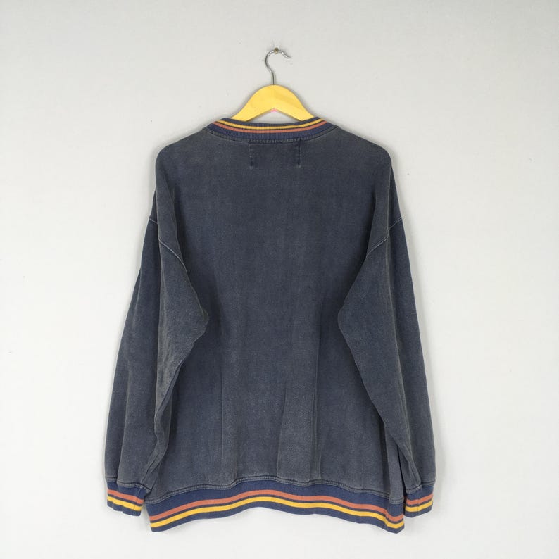 Puede incluir: Una sudadera azul oscuro con cuello redondo y mangas largas, colgada de una percha amarilla. El cuello y el dobladillo presentan rayas amarillas, naranjas y azules. La prenda tiene un aspecto descolorido y vintage.
