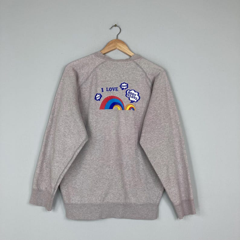 May include: A heather gray sweatshirt with a crew neck and long sleeves. The back features a colorful rainbow design with the text "I LOVE BODY GLOVE" in blue and white bubble letters. The sweatshirt has a ribbed hem and cuffs.