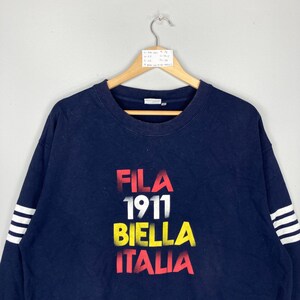 May include: Navy blue sweatshirt with the text "FILA 1911 BIELLA ITALIA" in red, yellow, and white. The sweatshirt has white stripes on the sleeves. The garment is hanging on a wooden hanger.