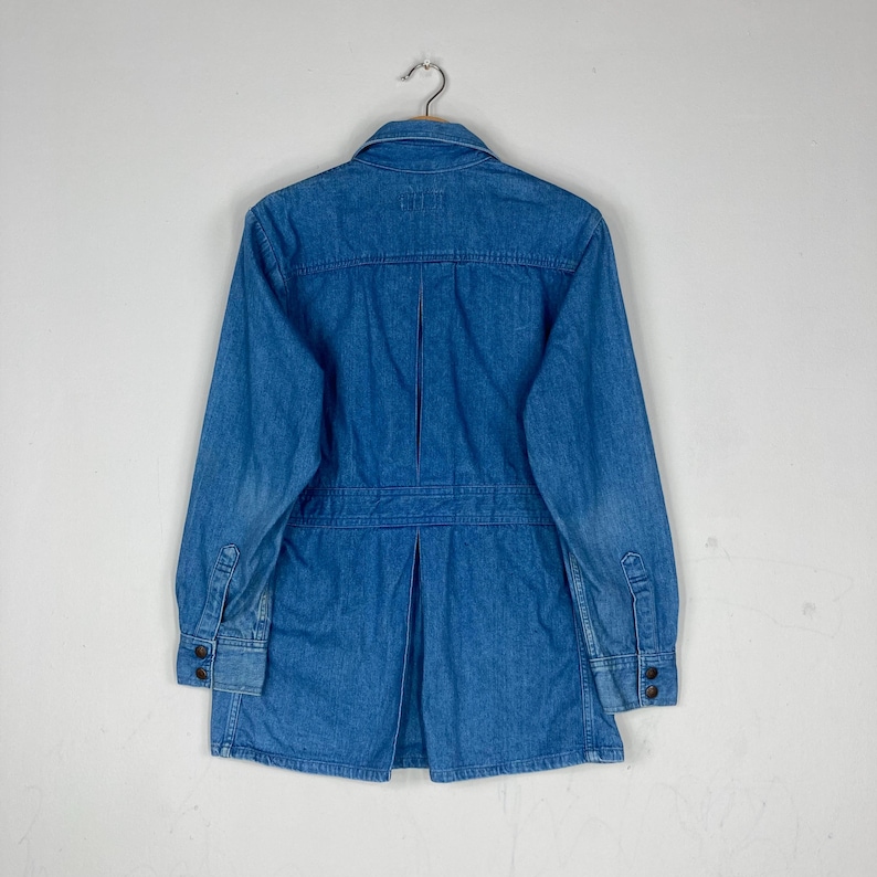 K&ouml;nnte beinhalten: Hellblaue Jeansjacke mit langen &Auml;rmeln, gekn&ouml;pften Manschetten und plissiertem R&uuml;cken. Die Jacke h&auml;ngt an einem Metallb&uuml;gel vor einem schlichten wei&szlig;en Hintergrund. Die Jacke hat einen klassischen Kragen und eine Knopfleiste.