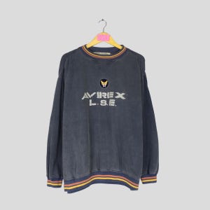 Puede incluir: Sudadera vintage azul marino con el texto "AVIREX L.S.E." en blanco y un peque&ntilde;o emblema encima. El cuello y los pu&ntilde;os tienen detalles a rayas en amarillo, rojo y azul. La sudadera est&aacute; colgada de una percha de madera.