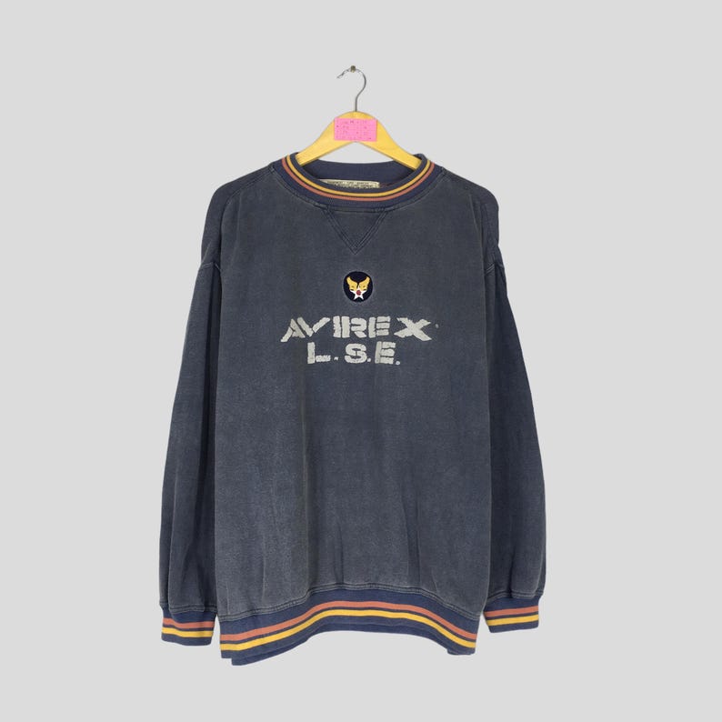 Puede incluir: Sudadera vintage azul marino con el texto "AVIREX L.S.E." en blanco y un peque&ntilde;o emblema encima. El cuello y los pu&ntilde;os tienen detalles a rayas en amarillo, rojo y azul. La sudadera est&aacute; colgada de una percha de madera.