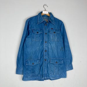 K&ouml;nnte beinhalten: Vintage blaue Jeansjacke mit vier Vordertaschen und Knopfverschluss. Die lang&auml;rmlige Jacke hat einen Kragen und ein kleines rotes Logo auf der Brust. Die Jacke h&auml;ngt an einem Kleiderb&uuml;gel.