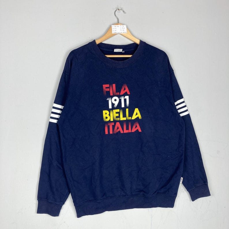 May include: Navy blue sweatshirt with the text "FILA 1911 BIELLA ITALIA" in red, white, and yellow. The long sleeves have white stripes near the cuffs. The sweatshirt is hanging on a wooden hanger.
