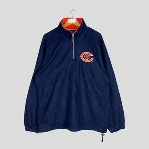 Puede incluir: Jersey de forro polar azul marino con el logotipo de los Chicago Bears en el pecho. El cuello es naranja y la cremallera es plateada. La parte superior de manga larga tiene un cordón en la parte inferior.