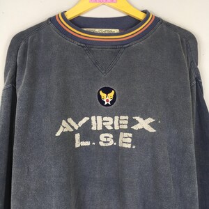 Puede incluir: Una sudadera azul descolorida con el nombre de la marca "AVIREX L.S.E." en letras blancas. El cuello redondo tiene un ribete a rayas azules, naranjas y amarillas. Un peque&ntilde;o emblema est&aacute; centrado encima de la marca.