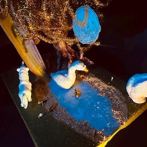 Könnte beinhalten: Ein Miniatur-Diorama mit einem Teich mit einer weißen Gans und einem kleinen Boot. Im Hintergrund ein Baum mit goldenem Lametta und einem blauen, cartoonartigen Ornament. Die Szene ist auf einer grünen und braunen Basis aufgebaut.