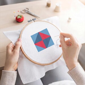 May include: A cross-stitch project in progress, featuring a geometric design in a wooden embroidery hoop. The design includes red and blue triangles on a white fabric. A needle and thread are visible, along with a tomato-shaped pin cushion and scissors.